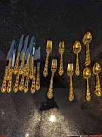 Full view of various gold-tone flatware items including knives, forks, and spoons arranged on dark surface displaying handles and blades/bowls.