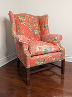 Front view of orange floral bird pattern wingback chair with solid wood legs on hardwood floor against beige wall.