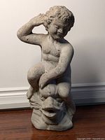 Front view of concrete statue showing cherub sitting on fish with mouth open for water spout, visible wear and some cracks.