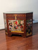 Angled front view showing detailed hand-painted scenes of Burmese people in traditional clothing with a red/maroon base and gold accents. Glass top visible with slight corner damage.