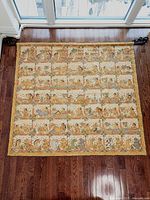 Full view of the large square tapestry laid flat on wood floor showing detailed hand-painted scenes in a grid format and decorative border.