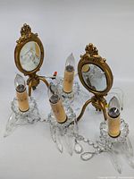 Pair of ornate brass and crystal two-bulb wall sconces with oval mirrored backs and candle-shaped bulbs. Includes crystal chains and prisms. Photo taken to show overall design and condition.