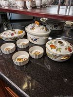 Photo showing entire set of seven Royal Worcester Evesham porcelain pieces laid out on black countertop, includes all casserole dishes and bowls with fruit designs and gold details.