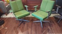 Two matching green mid-century modern swivel chairs with wood arm accents seen from the front and slightly angled side view on wood flooring with additional furniture in the background.