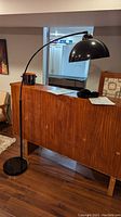 Full view of black arch lamp curved over a wooden cabinet in living room with black dome shade, foot switch visible on cord