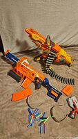 Large and medium size Nerf blasters with ammunition belt and magazines, foam darts, and glasses spread on fabric background.