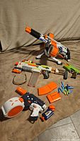 Three Nerf blasters with multiple orange and translucent magazines, darts, and safety glasses on a brown couch.