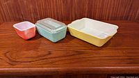 Three Pyrex dishes displayed side by side on wooden surface against wood wall: small coral dish, medium turquoise dish with lid, large yellow dish.