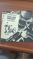 Front cover of 'Lace and Whiskey' vinyl album by Alice Cooper showing title, artwork with revolver and shot glass.