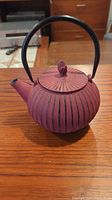 Red cast iron teapot shown from front on wooden table, highlighting ribbed design and lid finial.