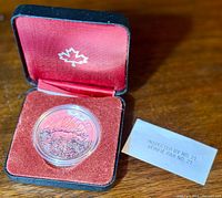 Opened display case showing the silver coin in a clear capsule on a red velvet cushion, with an inspection slip beside it.