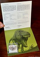 Coin housed in protective sleeve laid on green folder featuring large detailed T. rex illustration and certificate of authenticity text