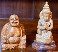 Two carved wooden sculptures side by side on a wooden surface with a wooden background. Left figure is a laughing Buddha, right figure a finely detailed seated figure on a base.