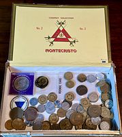 Top view of open Montecristo cigar box revealing a diverse assortment of coins of various currencies and conditions, arranged loosely inside.