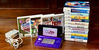 Photo of Nintendo 3DS console in purple with charger and 13 game cases arranged next to it.
