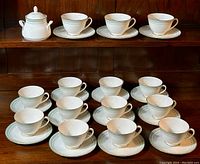 Overview of 14 tea cups, 14 saucers and one lidded sugar bowl arranged on a wooden shelf