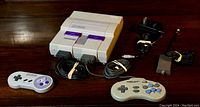 Super Nintendo console front view showing two controllers and cables laid out, including power adapter and AV cable
