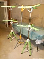 View of two green/white/silver two-tier adjustable drying racks with collapsible arms and tripod bases, one showing two wheels and one showing slightly damaged wheels, standing in a basement area.