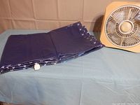 Photo showing the folded navy blue Intex air mattress with full velvet bottom and a white valve, placed next to the yellow-beige Marvin Studio 12" blade fan.