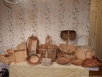 Wide view showing assorted baskets in different shapes, sizes, and materials arranged on a table against a floral wall background. Includes rectangular, oval, and round baskets with single, double and no handles.