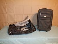 Two black bags on light blue surface against beige backdrop: lying black Samsonite duffle bag and upright black Ascot rolling carry-on suitcase.