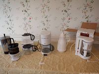 Photo showing multiple electric drip coffee makers, a vintage metal percolator, a white ceramic coffee thermos, reusable coffee filters, and glass jars all arranged on a table with floral wallpaper background.