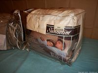 Cream-colored queen-sized Sunbeam electric blanket packaged in clear plastic with dual controls and visible cords.
