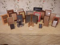 Photo showing 22 assorted photo frames arranged on a table against floral wallpaper background.