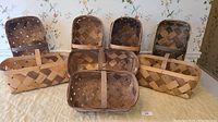 Photo of 8 woven fruit baskets arranged in two rows. Seven baskets are larger rectangles with handles, one smaller basket is front and center. Natural wood tone variations, visible dust, and light use.