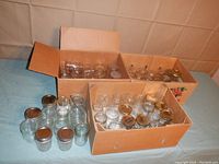 Three boxes filled with clear glass jelly size jars and some jars placed outside boxes on surface. Boxes open showing mixed jars sizes and some with lids.