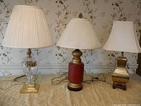 Three lamps on table against floral wallpaper, showing glass and gold lamp with pleated ivory shade on left, red and black ceramic lamp with pleated ivory shade center, and oriental metal lamp with white shade right