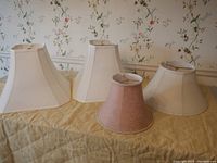 Four lamp shades displayed on a yellow-covered table with floral wallpaper background: two off-white octagon shapes, one pink damask flared bell, and one ivory flared bell shade.