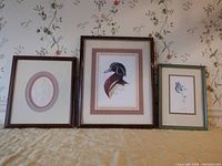 Three framed nature prints displayed side by side: an embossed butterfly print with double oval mats in brown wood frame, a detailed wood duck print with double mats in brown burled plastic frame, and an embossed iris print by Dot Goodwin with double oval mats in beveled brown wood frame.
