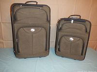 Photo showing the exterior front view of the two-piece American Tourister luggage set in olive green and black, with the larger bag on the left and smaller bag on the right.