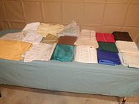 Wide angle photo showing the full assortment of folded pillowcases and flat sheets arranged on a table, featuring a variety of colors including mustard yellow, teal, cream floral, solid white with burn hole, red, green, and brown.