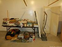 Wide view showing fabric piles on table, boxes below, foam forms rolled vertically at right, ironing boards, vintage Jiffy steamer, and wooden trifold display in background.