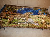Full view of vintage velvet wall tapestry featuring a safari hunt scene with tiger and antelope, placed on a table showing blue fringe on sides and rich colorful design.
