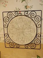 Square metal wall hanging frame containing circular ornate resin medallion with floral and leaf design, sitting on floor near wallpapered wall.