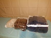 Three warming throws in clear storage bags placed side-by-side on a surface with a tan background.