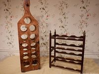 Photo showing both wine racks together on table with floral wallpaper background: tall oak colored rack with 11 circular bottle holes, smaller cherry rack with wave-shaped shelves and acorn finials