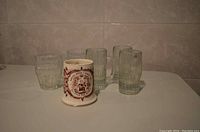 Set of three tall glass mugs, two short glass mugs, and one smaller ceramic mug arranged on a white surface against a tiled wall background.