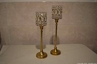 Two gold-tone metal candle holders with glass rhinestone decoration, one taller than the other, placed on a light-colored surface against a beige background.