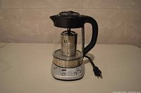 Full view of the electric tea brewer showing handle, brewing basket, water reservoir, and power cord.