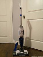 Full front view of the upright Hoover battery-powered vacuum mop showing the handle, upper body with Hoover logo, transparent water tank section and base on wood floor near white door.
