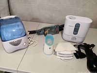 Photo showing Boneco ultrasonic humidifier, Vicks humidifier, Vicks portable steam inhaler, Epi Sauna facial steamer, Ab Isolator VHS tape, and posture strap on a white surface.