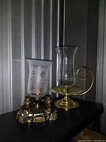 Photo showing brass electric lamp with owl figures and floral frosted glass shade next to brass candle holder with glass hurricane shade