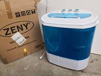 Zeny washing machine next to its original box, showing front and side of the unit with blue tubs and control knobs on top.