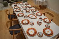 Entire dinnerware set displayed on a white tablecloth with wooden chairs around the table under indoor lighting, showcasing the general layout and quantity of items including plates, cups, serving pieces.