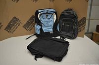 Three backpacks shown, blue and white Heys ePac backpack at front left with black base and sides, green backpack at back right with bright green zipper accents, black backpack at front right partially visible.