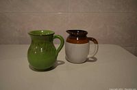 Two clay pitchers displayed side by side, one green and glossy, the other beige with brown top.